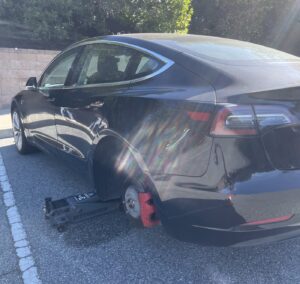 Axl Roadside flat tire job on a Tesla in a parking lot: rear wheel off, red Tesla brake caliper showing, car on a floor jack