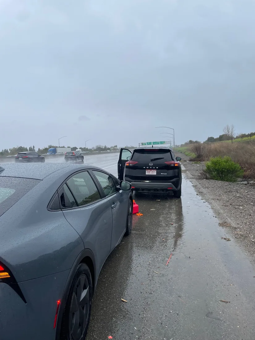 A sedan pulled over on a rain-soaked shoulder with its door open and a cone set out behind it.